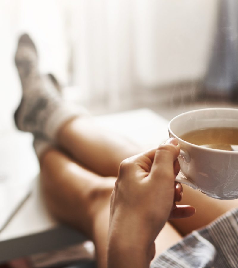 Cup of tea and chill. Woman lying on couch, holding legs on coffee table, drinking hot coffee and enjoying morning, being in dreamy and relaxed mood. Girl in oversized shirt takes break at home.